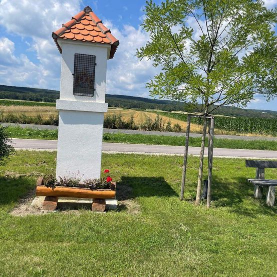 Eine kleine weiße Struktur mit einem roten Dach steht in einem Grasfeld. Es hat ein Fenster mit einem Metallgitter und einen Blumenkasten mit Blumen davor. Ein Baum und eine Bank sind in der Nähe.