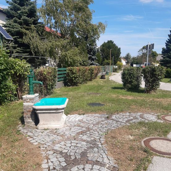 Ein kleiner Steinbrunnen mit einer grünen Pumpe befindet sich in einem Grasbereich, umgeben von Büschen und Bäumen. Ein Solarpanel ist links an einem Pfosten montiert.