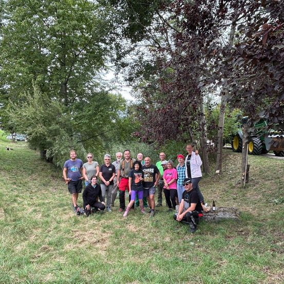 Eine Gruppe von Menschen posiert für ein Foto in einem Grasbereich mit Bäumen und einem Traktor im Hintergrund.
