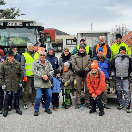Eine Gruppe von Menschen, einschließlich Erwachsener und Kinder, steht vor Traktoren und Gebäuden und trägt Winterkleidung und teilweise Mützen. Sie scheinen für ein Foto zu posieren.