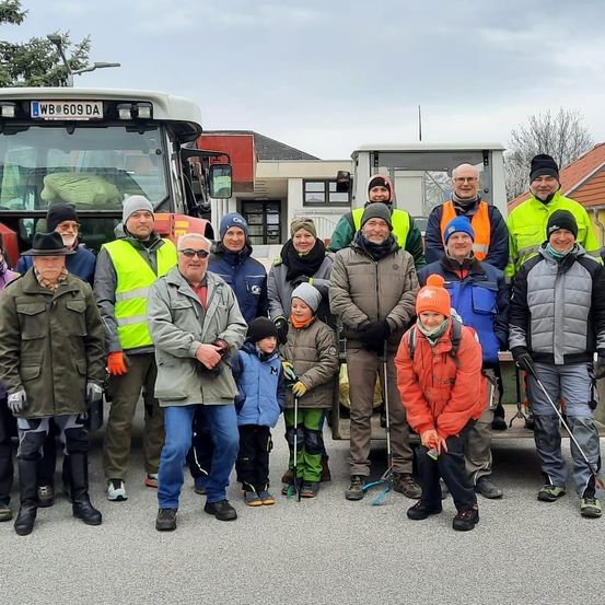 Eine Gruppe von Menschen, einschließlich Erwachsener und Kinder, steht vor Traktoren und Gebäuden und trägt Winterkleidung und teilweise Mützen. Sie scheinen für ein Foto zu posieren.