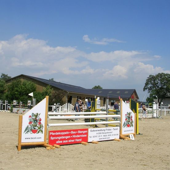 Ein Pferdesprungparcours ist in einem sandigen Bereich aufgebaut. Es gibt verschiedene Banner mit Text und Logos. Im Hintergrund befindet sich ein Gebäude mit Solarpanelen und mehrere Personen stehen herum. Der Himmel ist blau mit einigen Wolken.
