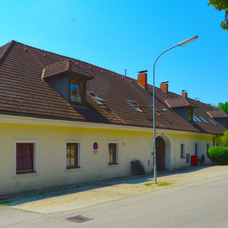 Eine Reihe gelber Häuser mit braunen Dächern, mehreren Fenstern und einer Straßenlaterne. Ein Mülleimer und einige Pflanzen sind davor.