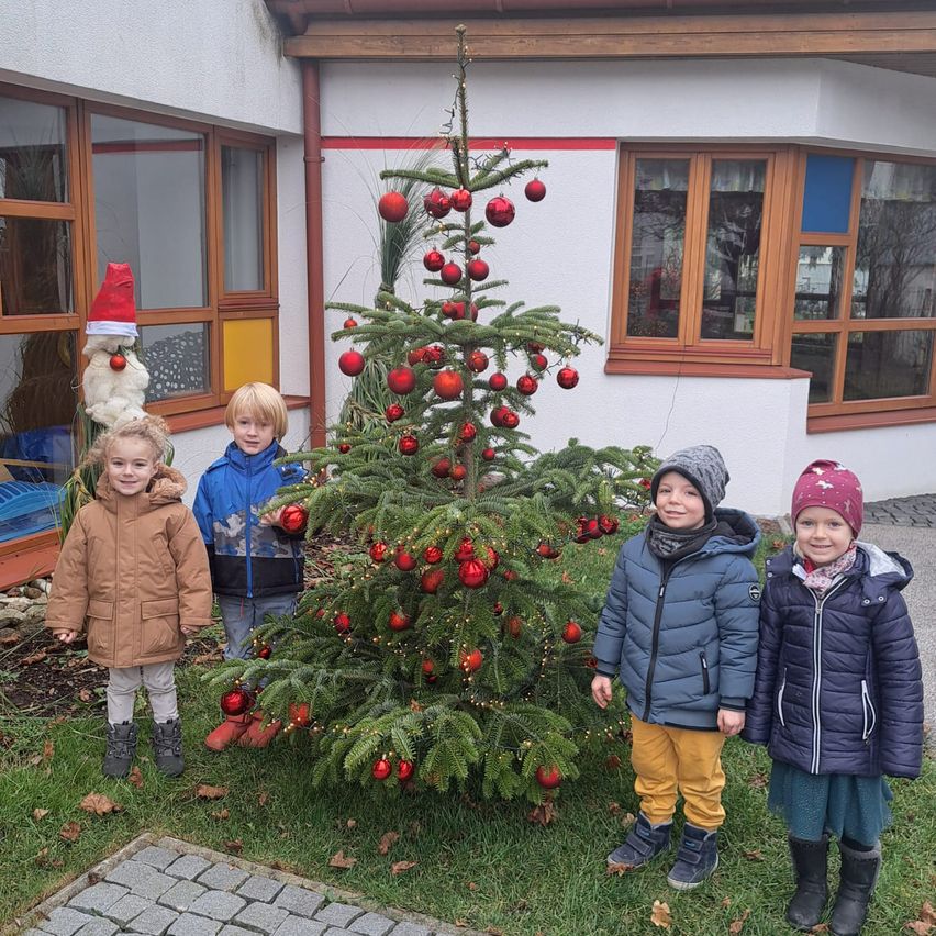 Vier Kinder stehen um einen Weihnachtsbaum mit roten Kugeln vor einem Gebäude mit Glasfenstern.