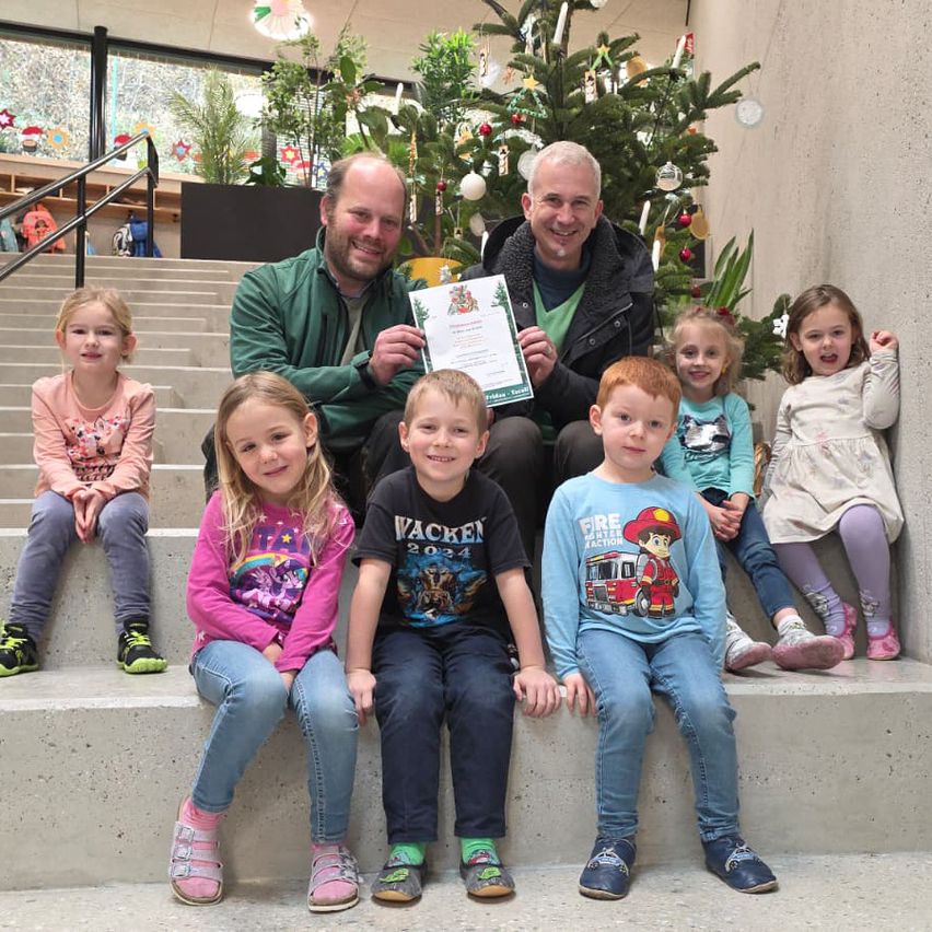 Mehrere Erwachsene und Kinder sitzen auf Stufen mit Weihnachtsbäumen im Hintergrund. Zwei Männer halten ein Zertifikat.