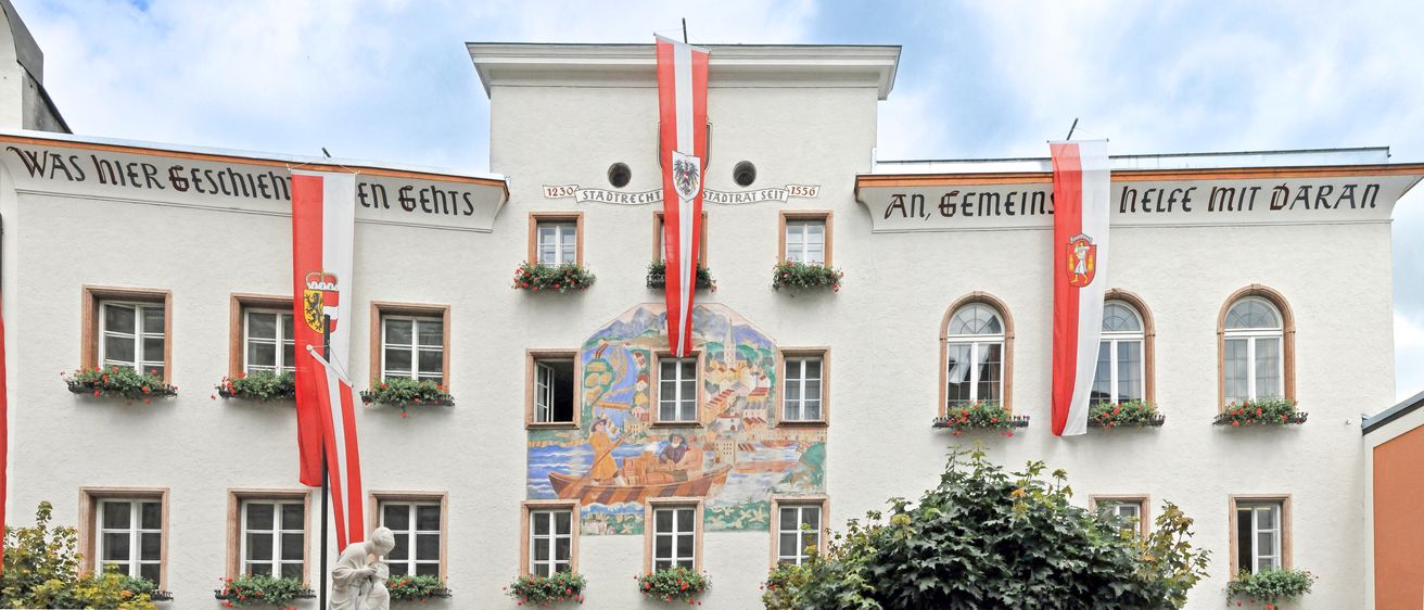 Ein weißes Gebäude mit einer rot-weißen Flagge, einem Wandbild und historischen Texten. Es hat Fenster, die mit Blumenkästen geschmückt sind.