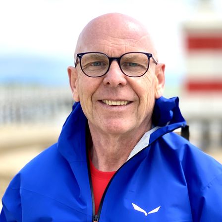 Ein älterer Mann mit kahlem Kopf, der eine blaue Jacke mit einem weißen Logo und einem roten Unterhemd trägt, lächelt vor einem verschwommenen Hintergrund.