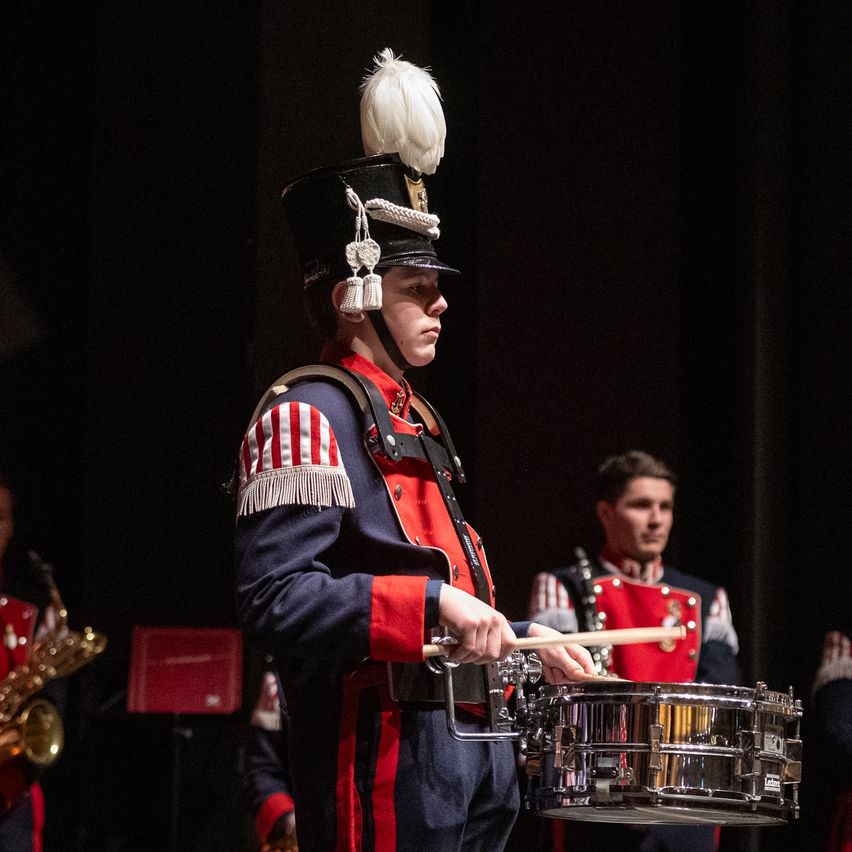 Ein Mitglied einer Marschkapelle spielt die Snare-Drum bei einer Aufführung. Er trägt ein militärisches Outfit mit einem roten und weißen Streifen auf dem Ärmel.