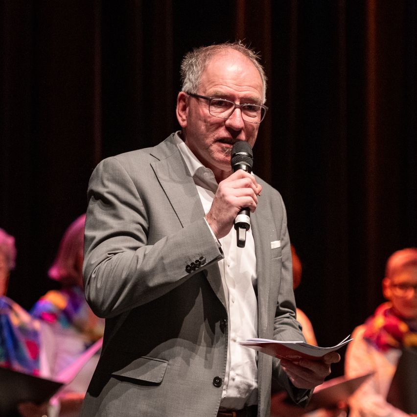 Ein Mann in einem grauen Anzug und mit Brille steht auf der Bühne, hält ein Mikrofon in der Hand. Er liest von einem Blatt Papier. Hinter ihm sitzen eine Gruppe von Menschen und lauschen.