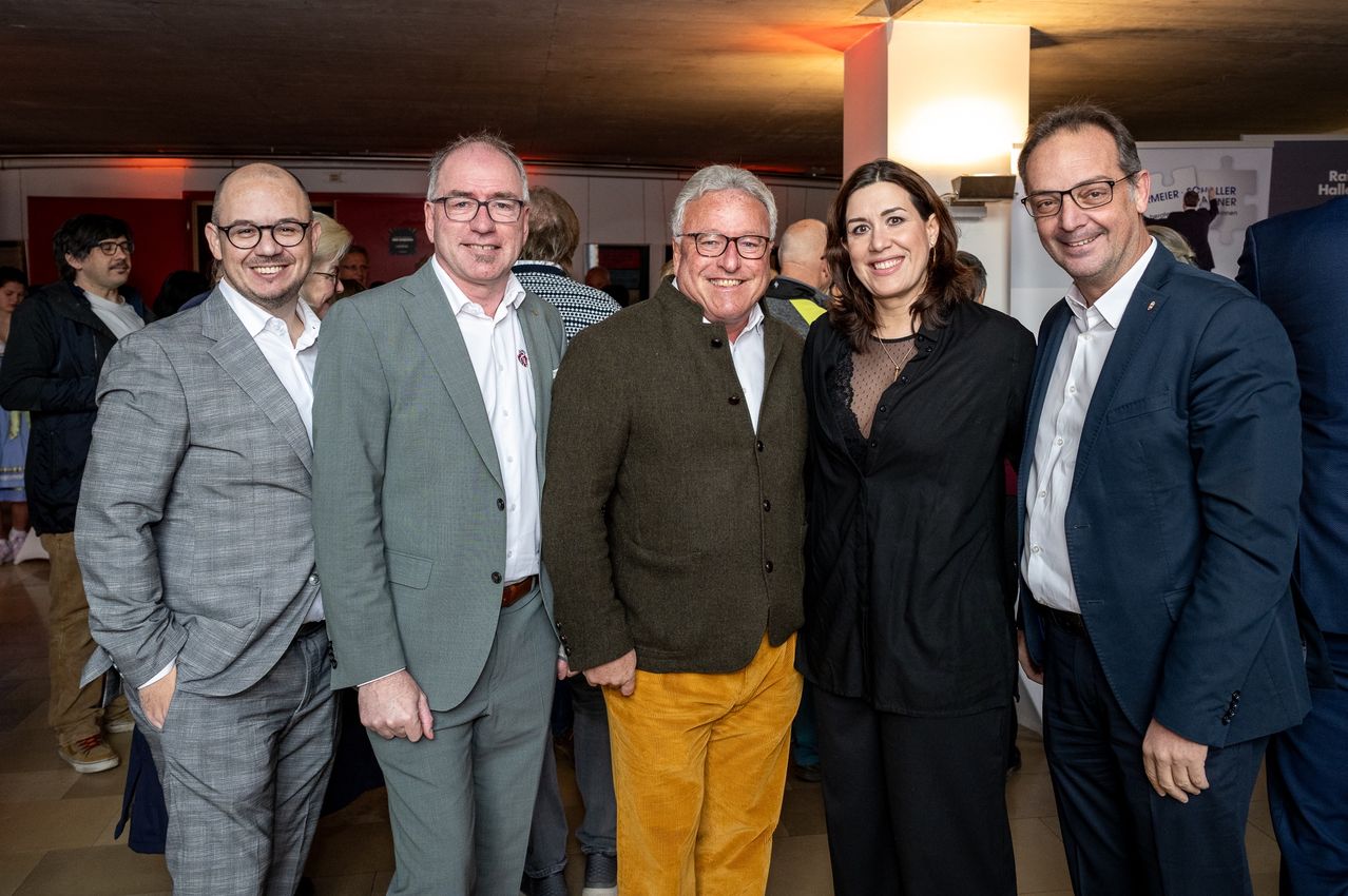 Fünf Erwachsene lächeln und posieren für ein Foto in einem Raum, wahrscheinlich bei einer Veranstaltung. Der Mann in der Mitte trägt gelbe Hosen.