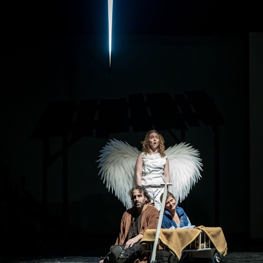 Eine Frau mit weißen Flügeln steht auf einer Bühne mit zwei Personen. Eine sitzt mit einem Schwert und die andere liegt auf einem Bett.
