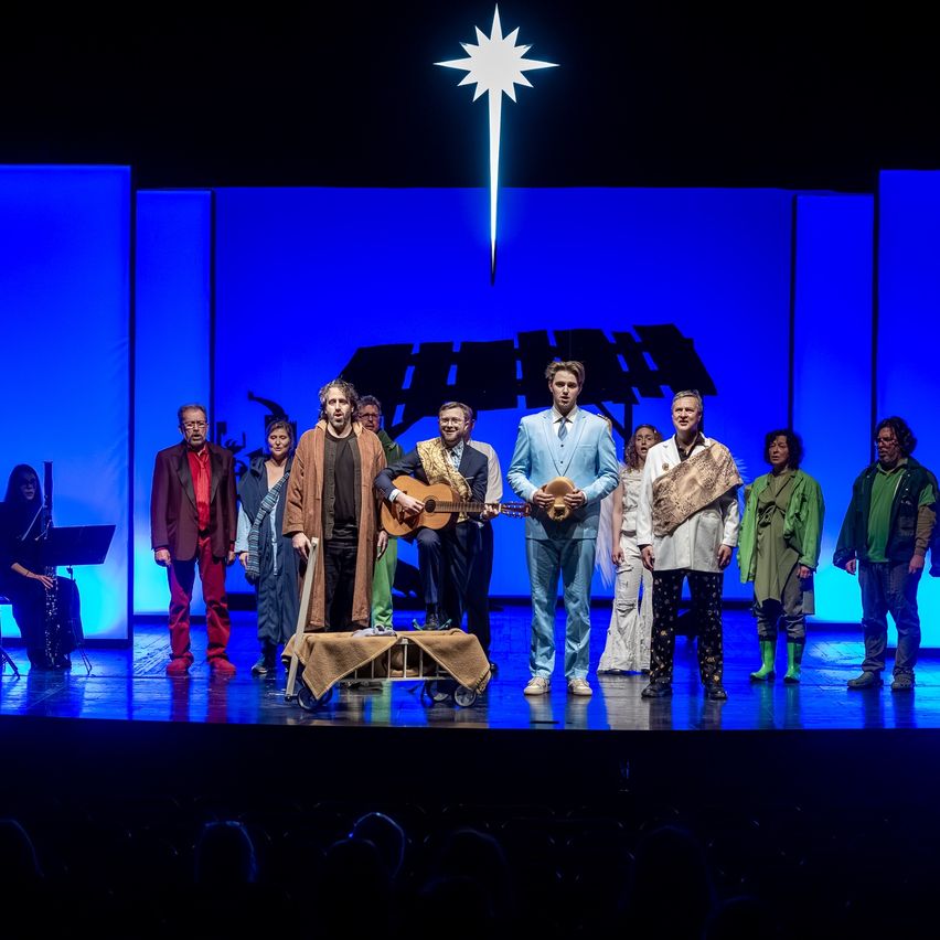 Eine Gruppe von Darstellern steht auf der Bühne, einer spielt Gitarre. Die Bühne ist blau beleuchtet und darüber befindet sich ein Sternsymbol.