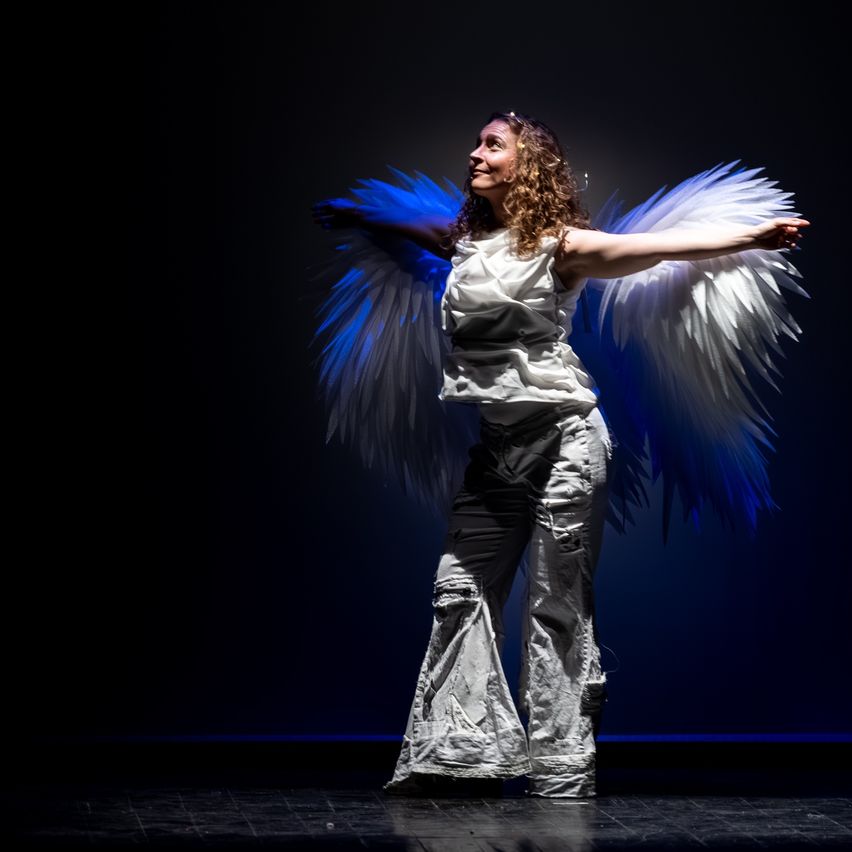 Eine Frau mit lockigem Haar und Flügeln tritt auf der Bühne auf, lächelt und streckt ihre Arme aus. Sie trägt ein weißes Kleid mit zerrissenen Hosen. Der Hintergrund ist dunkelblau.