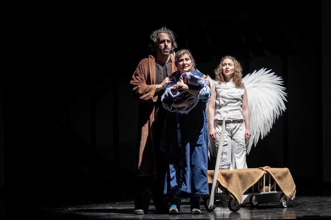 Drei Personen stehen auf der Bühne, eine hält ein Baby, eine trägt Engelsflügel und eine andere einen Umhang. Die Bühne ist dunkel.