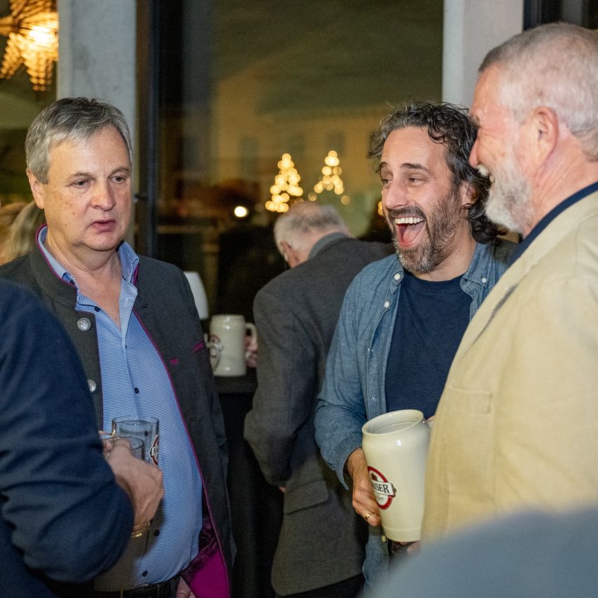 Eine Gruppe von Männern unterhält sich bei einer Indoor-Veranstaltung, einer hält einen Bierkrug mit dem Wort Berliner. Der Mann auf der linken Seite trägt eine Jacke, und der Mann auf der rechten Seite hat einen Bart.
