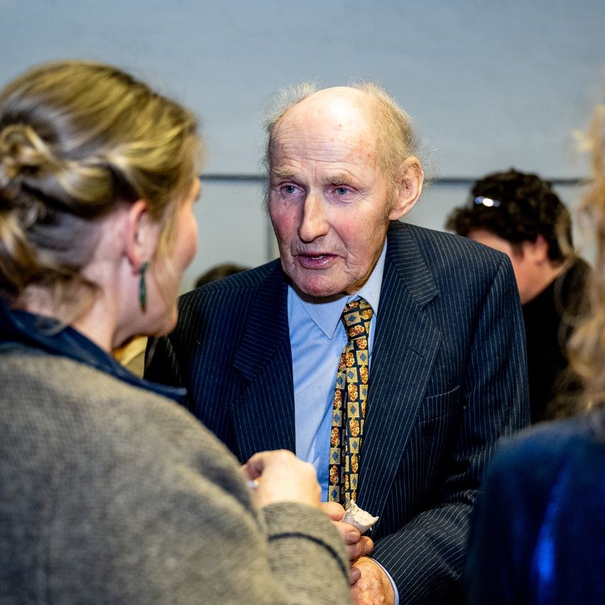 Ein älterer Mann in Anzug und Krawatte spricht bei einer Veranstaltung mit einer Frau. Er hält einen kleinen Gegenstand in der Hand.