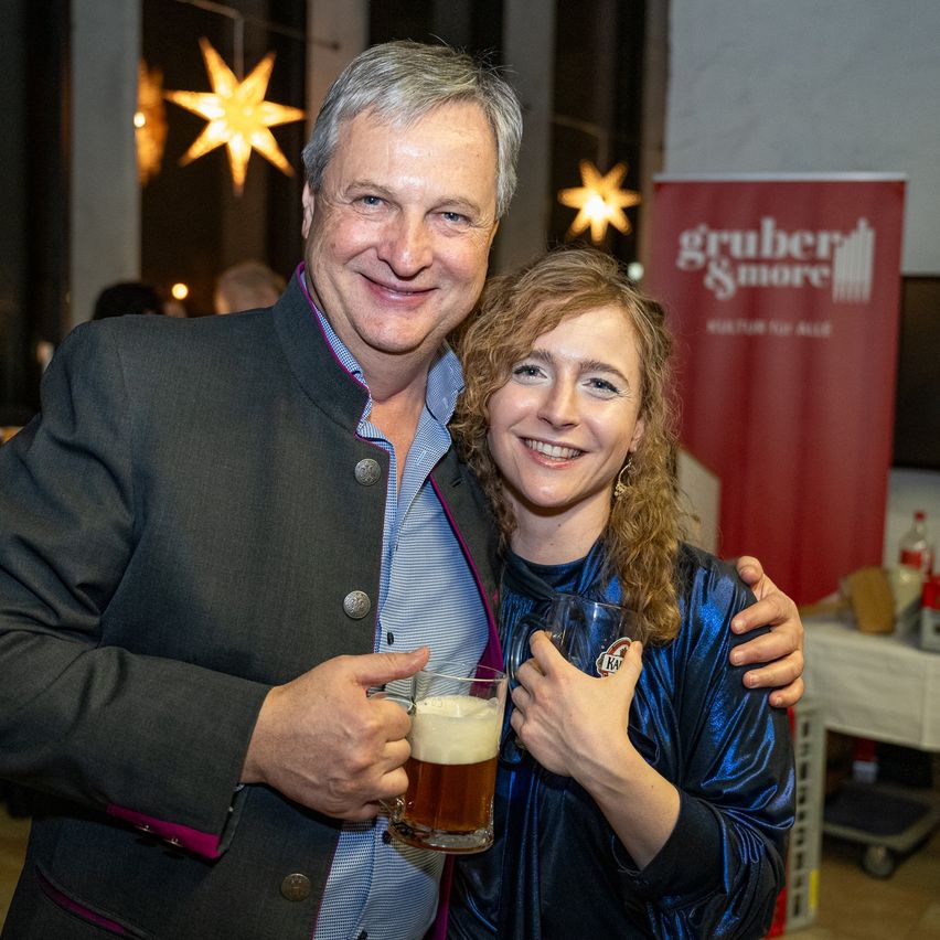 Ein Mann und eine Frau lächeln und halten Biergläser vor einem festlichen Hintergrund mit Sterndekorationen und einem roten Banner.