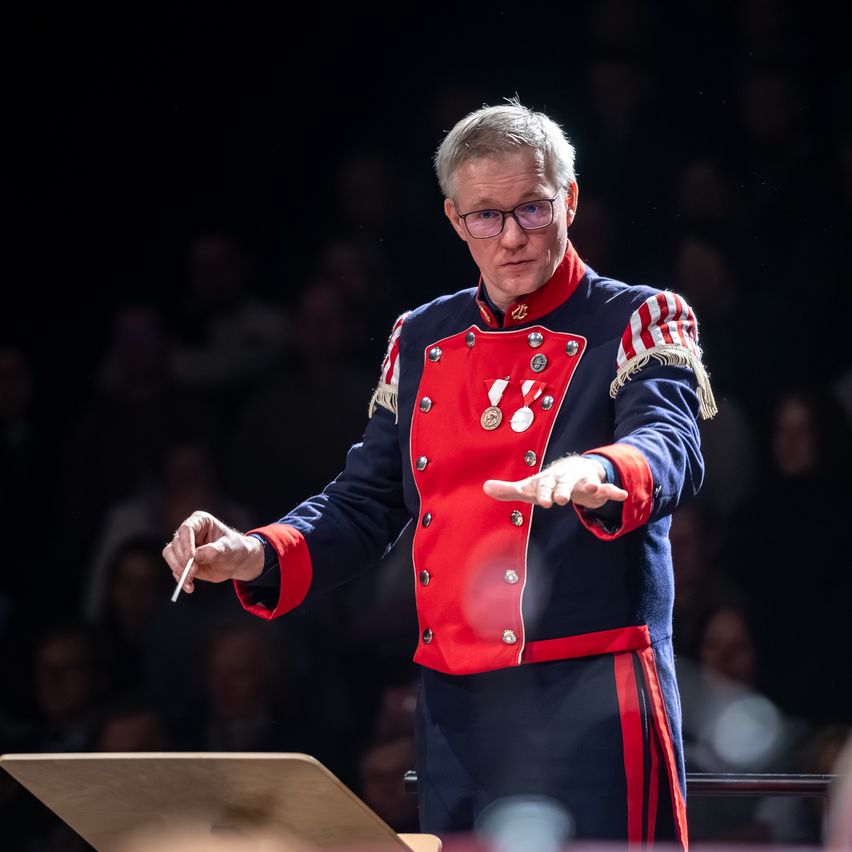 Ein Mann in rot-blauer Uniform dirigiert ein Orchester. Er hält einen Taktstock und steht vor einem Pult, während das Publikum zusieht.