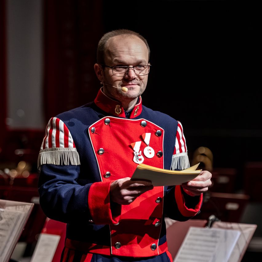 Ein Mann in einem roten und blauen Uniform mit Medaillen liest aus einem gelben Buch, während er ein Mikrofon hält.