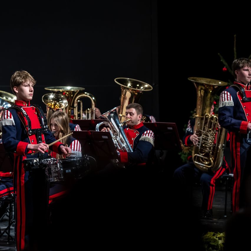 Eine Gruppe von Musikern in roten und blauen Uniformen spielt auf der Bühne mit verschiedenen Instrumenten, darunter Tubas und Trommeln. Ein junger Junge führt die Band an den Trommeln an.