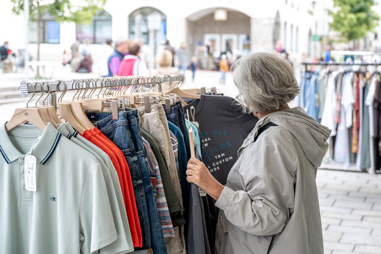 Eine ältere Frau betrachtet Kleidung in einem belebten Outdoor-Markt. Sie hält einen Kleiderbügel mit Kleidung in der Hand.