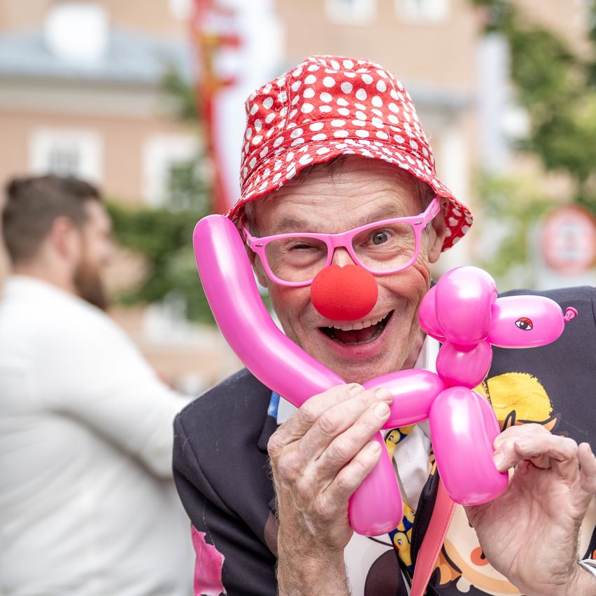 Ein Clown mit roter Clownsnase und Luftballon-Tier lächelt in die Kamera. Er trägt einen Hut mit roten Punkten und rosa Brille. Eine weitere Person steht verschwommen im Hintergrund.