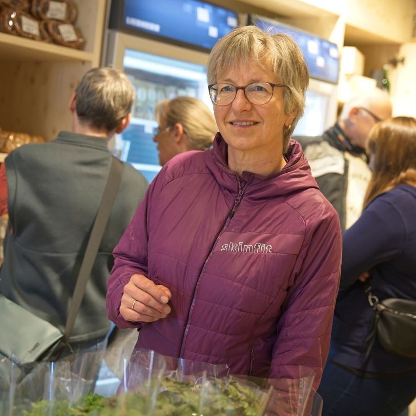 Eine Frau in einer lila Jacke lächelt, während sie einen Strauß grüner Kräuter in einem Geschäft hält. Hinter ihr stehen Leute, darunter ein Mann mit Brille.