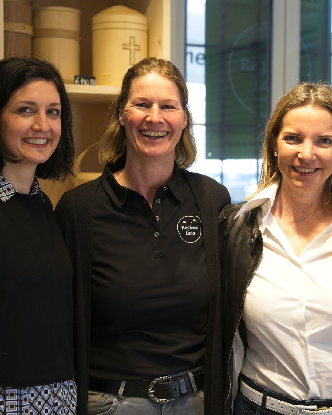 Drei Frauen lächeln und posieren für ein Foto. Die Frau in der Mitte trägt ein schwarzes Poloshirt mit einem Logo auf der linken Brust. Die anderen beiden Frauen tragen schwarze und weiße Kleidung. Dahinter steht ein Regal mit einem Kreuz und einem Schild.