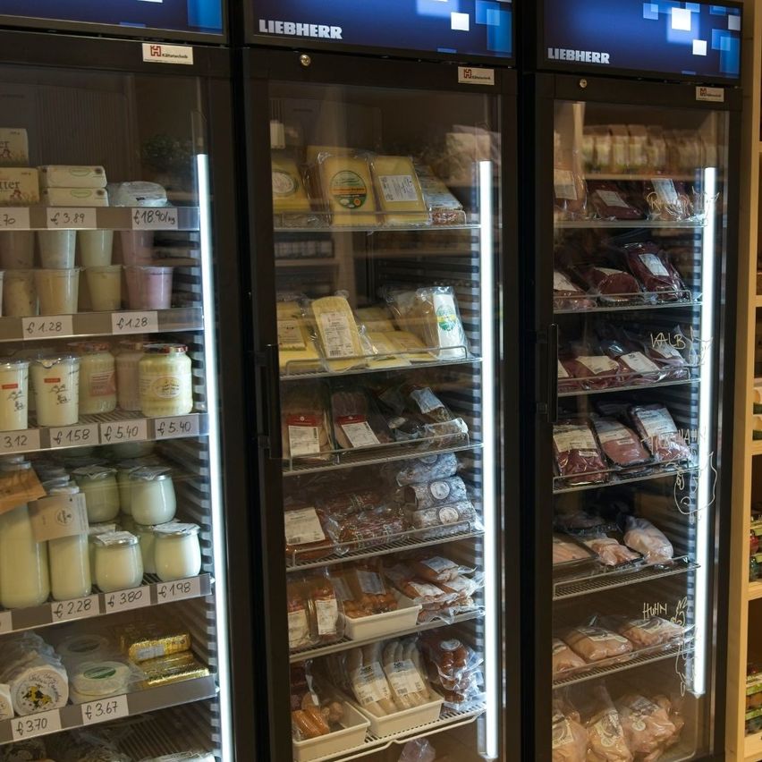 Zwei schwarze Kühlschränke zeigen eine Auswahl an Milch- und Fleischprodukten. Der linke Schrank enthält Milch- und Joghurtbehälter, während der rechte Schrank mit verpacktem Fleisch gefüllt ist. Oben auf den Schränken steht das Wort 'LIEBHERR'.