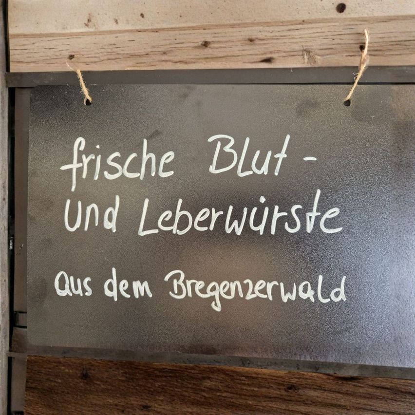 Ein Metallschild hängt an einer Holzwand und zeigt eine Botschaft in weißer Schrift. Es steht: 'frische Blut - und Leberwurst aus dem Bregenzerwald'. Das Schild hat eine reflektierende Oberfläche und ist mit einem Faden befestigt.