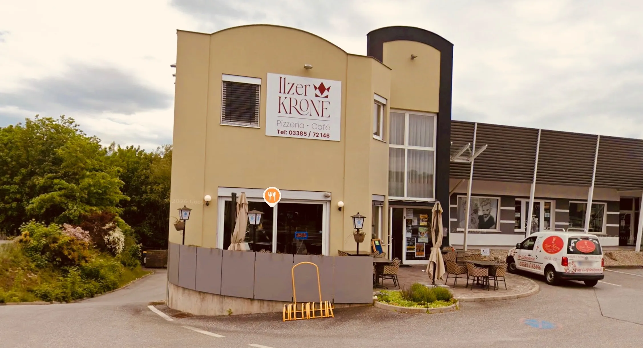 Die Iller Krone, eine Pizzeria und Café, zeigt ihren Namen auf einem weißen Schild über dem Eingang. Das Schild enthält auch die Telefonnummer 03385/72146. Außentische sind auf dem Bürgersteig aufgestellt, mit Schirmen für Schatten.