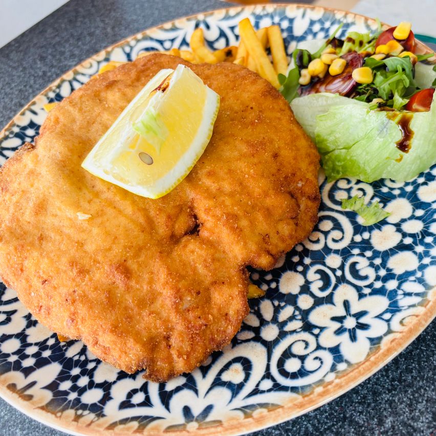 Ein Teller mit gebratenem Schnitzel, einer Zitronenscheibe, Pommes und Salat mit gemischtem Gemüse und Dressing.