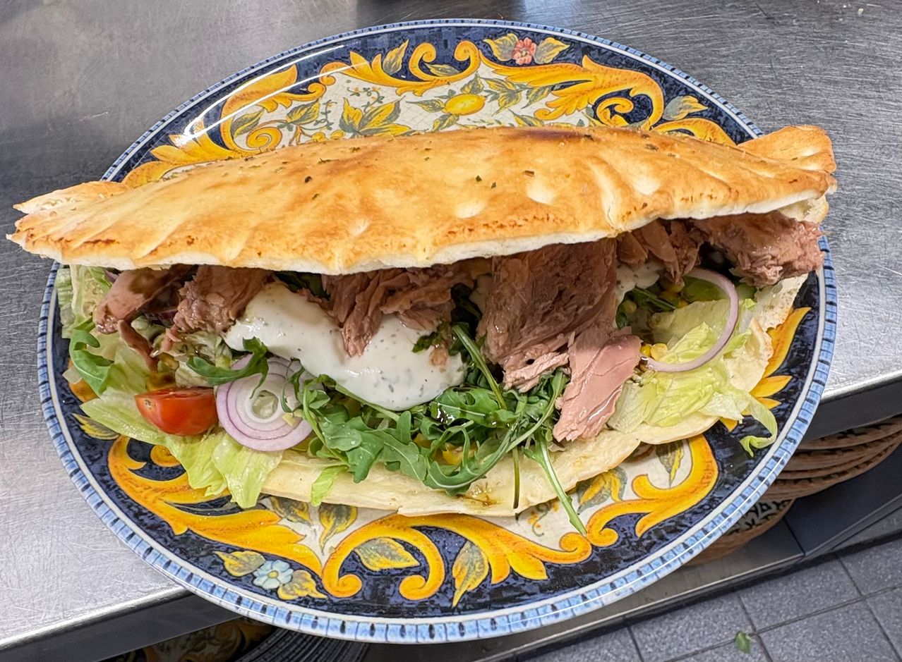 Ein Teller mit einem Sandwich, bestehend aus einem knusprigen Brötchen gefüllt mit dünn geschnittenem Fleisch, weißer Sauce, grünem Gemüse und roten Zwiebeln. Der Teller hat ein dekoratives Design.