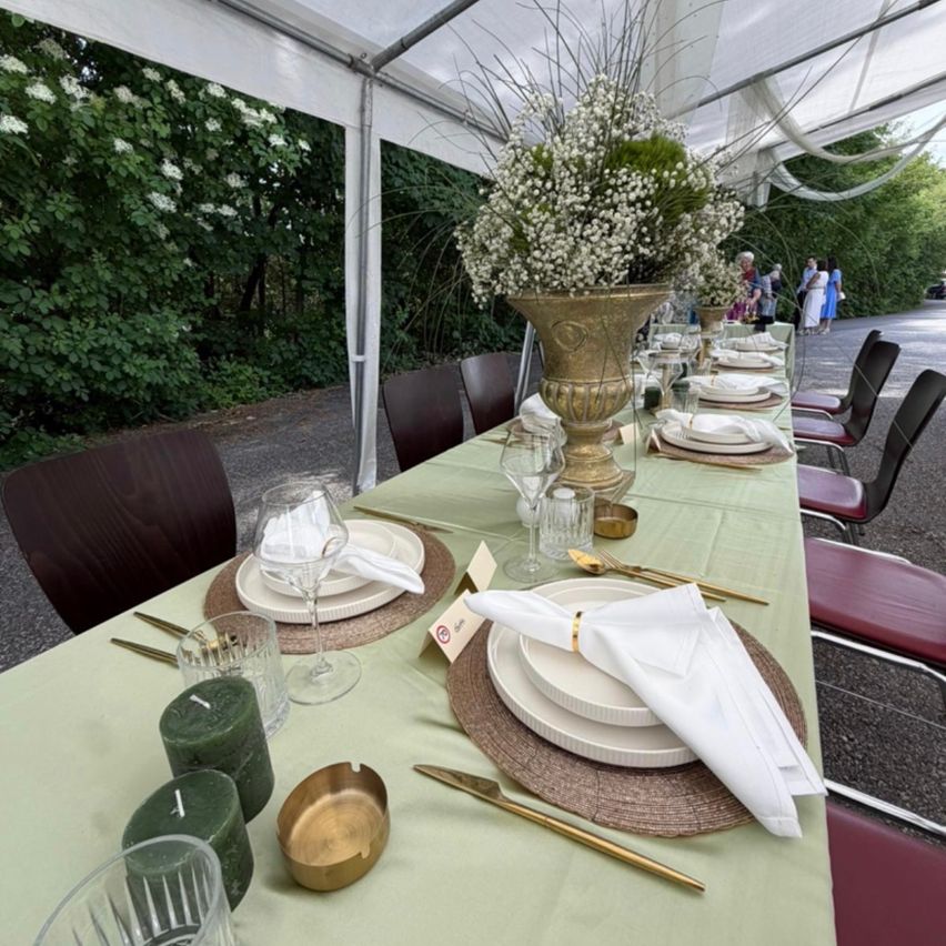 Ein langer Tisch für eine formelle Veranstaltung mit weißen Tellern, goldgeränderten Servietten und einem großen Blumenarrangement. Das Setting befindet sich unter einem weißen Zelt mit Grünpflanzen im Hintergrund.