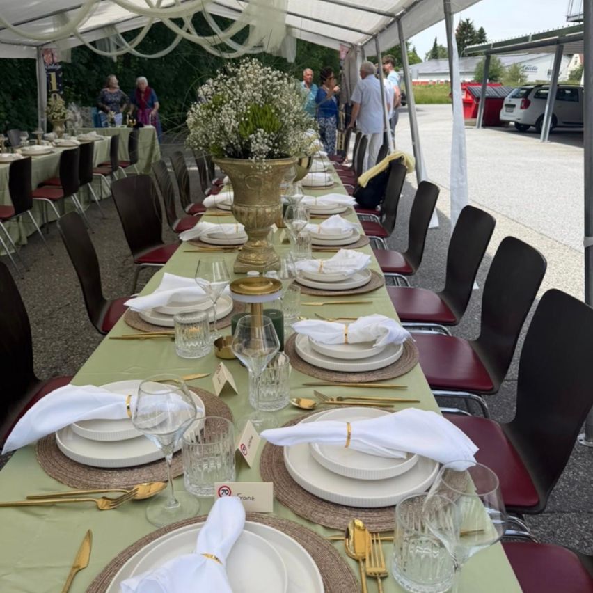 Ein Tisch für ein großes Ereignis im Freien, mit mehreren Tellern, Weingläsern und Besteck. Der Tisch ist mit einer grünen Tischdecke bedeckt, und die Stühle sind rot mit goldenen Akzenten.