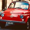 Ein leuchtend rotes Fiat 500 auf der Straße geparkt, mit 'CASA1' in weißer Schrift unter dem vorderen Stoßfänger. Das Auto hat klassische Merkmale und steht neben einem Gebäude.