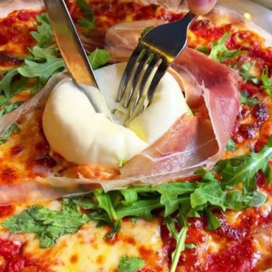 Eine Pizza, bei der ein Stück mit Messer und Gabel geschnitten wird, belegt mit Käse, Prosciutto und Rucola.