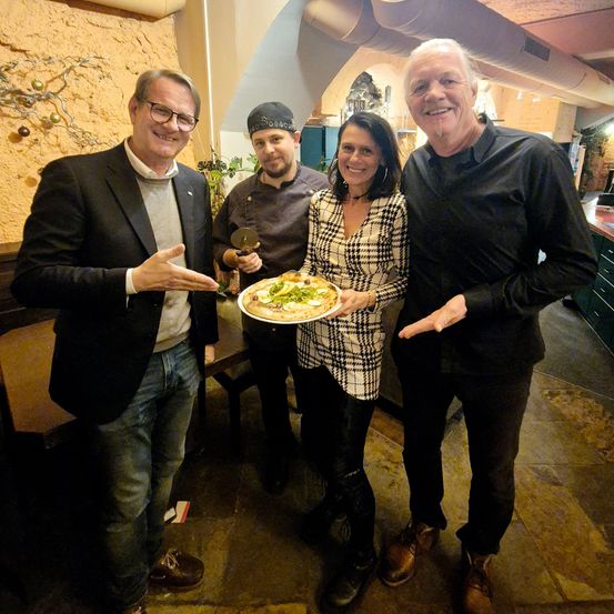 Vier Personen posieren für ein Foto in einem Restaurant und halten einen Teller mit Pizza.