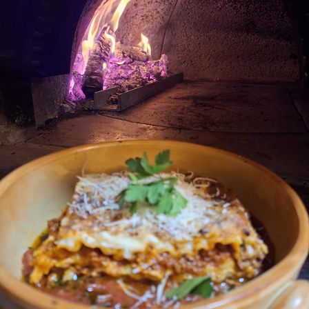Eine Schüssel Lasagne vor einem Ofen mit Feuer im Inneren. Das Gericht ist mit Käse und Petersilie belegt.