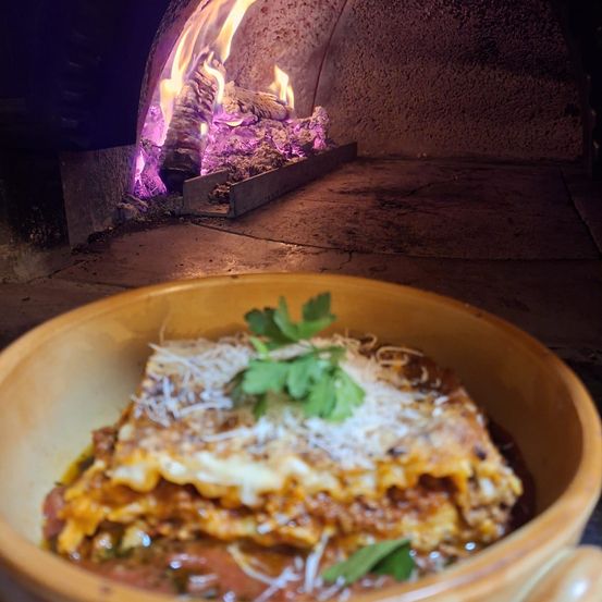 Eine Schüssel Lasagne vor einem Ofen mit Feuer im Inneren. Das Gericht ist mit Käse und Petersilie belegt.