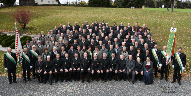 Eine große Gruppe von Menschen in Uniformen und Hüten posiert für ein Foto vor einem grünen Feld mit Büschen und Fahnen.