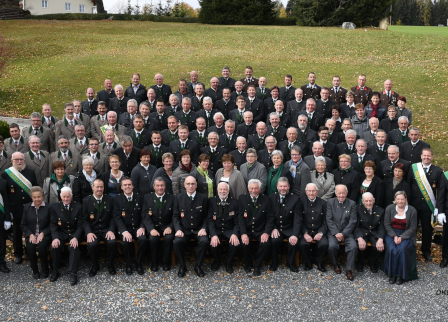 Eine große Gruppe von Menschen in Uniformen und Hüten posiert für ein Foto vor einem grünen Feld mit Büschen und Fahnen.