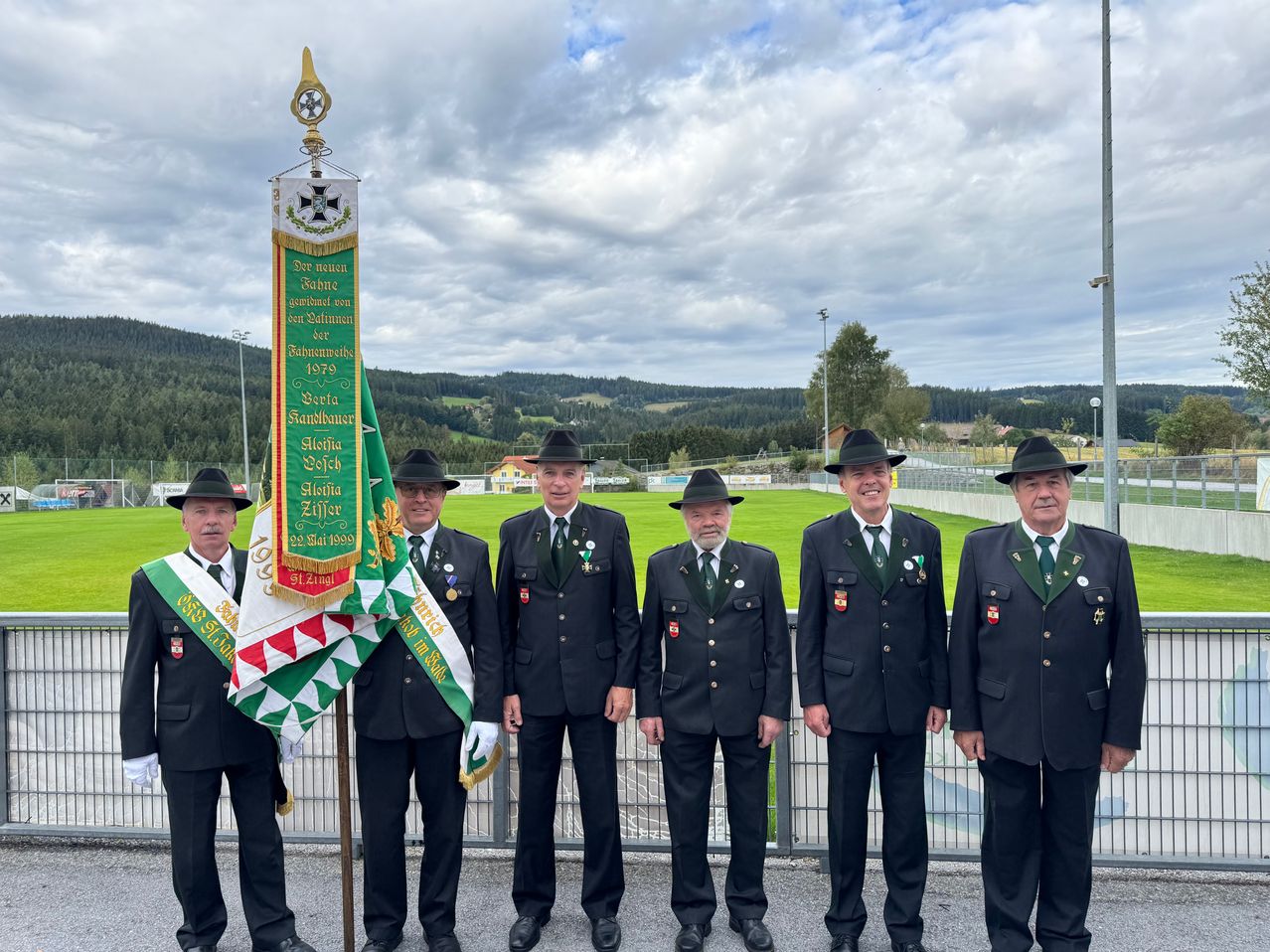 Fünf Männer in formellen Uniformen stehen vor einem Fahnenmast mit einem Banner, wahrscheinlich bei einer zeremoniellen Veranstaltung. Der Hintergrund zeigt ein Grasfeld und entfernte Berge.
