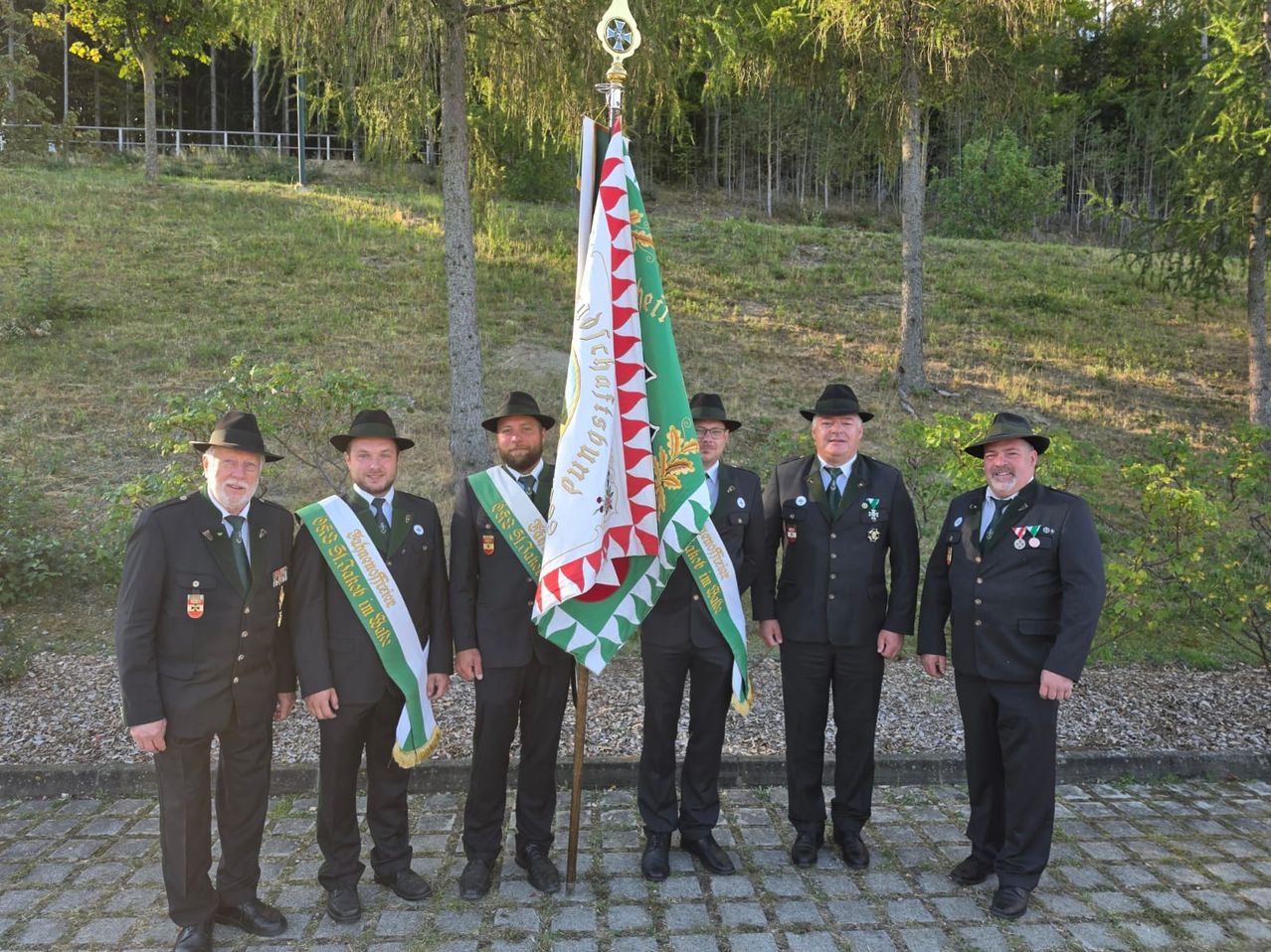 Fünf Männer in formellen Uniformen stehen vor einem Fahnenmast und halten eine Fahne mit rotem und weißem Design. Sie lächeln und scheinen für ein Foto zu posieren. Hinter ihnen befindet sich eine grasbewachsene Fläche mit Bäumen.