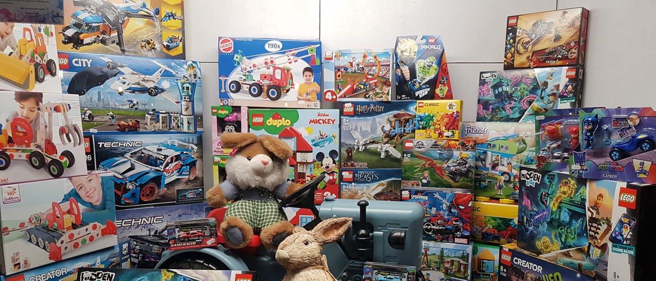 Eine Ausstellung verschiedener Spielzeugkisten an einer Wand, darunter LEGO-Sets, Micky-Maus-Spielzeug und Plüschtiere. Ein brauner Plüschhase sitzt auf einem Spielzeugtraktor mit einem großen Rad.