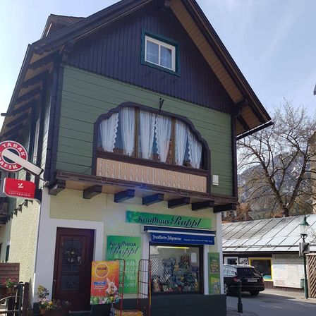 Ein grünes Geschäft namens Kaufhaus Rappl steht vor einem Berg. Das Geschäft hat weiße Vorhänge, einen Balkon und ein grünes Schild. Vor dem Geschäft ist ein schwarzes Auto geparkt.
