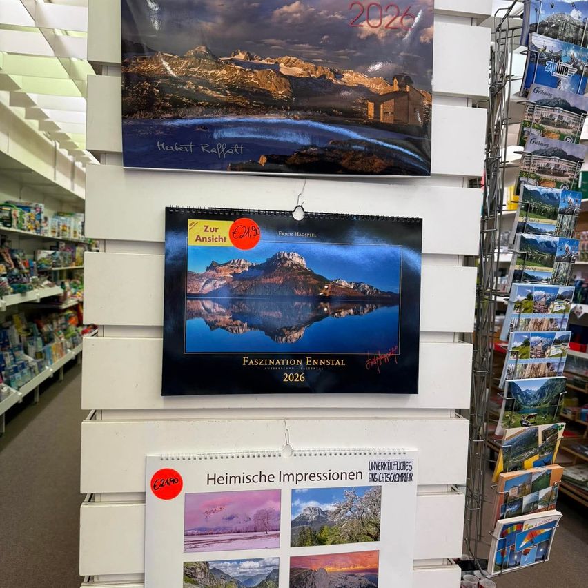 Ein Laden zeigt eine Wand voller Kalender. Einer ist für 2026 mit einem Berg und einem See, signiert von Herbert Raffalt. Ein weiterer Kalender zeigt Ennstal mit einer Reflexion im Wasser. Darunter befindet sich ein weiterer Kalender für 2026 mit Bergszenen, beschriftet mit 'Heimische Impressionen'.