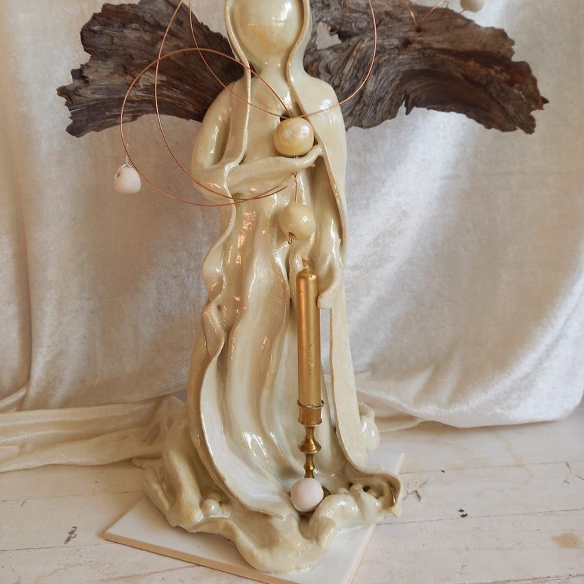 Eine Keramikstatue einer Frau mit einer Kerze steht auf einer weißen Basis. Sie trägt ein weißes Gewand und hat Holzflügel mit Perlen.