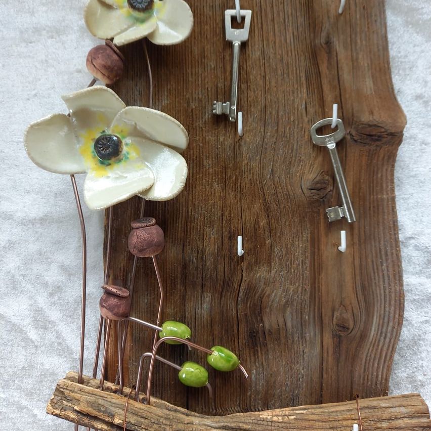 Ein Holzstück mit weißen Keramikblumen, Kupferstielen, grünen Perlen und zwei silbernen Schlüsseln. Das Holz ist gealtert und hat eine natürliche Textur.