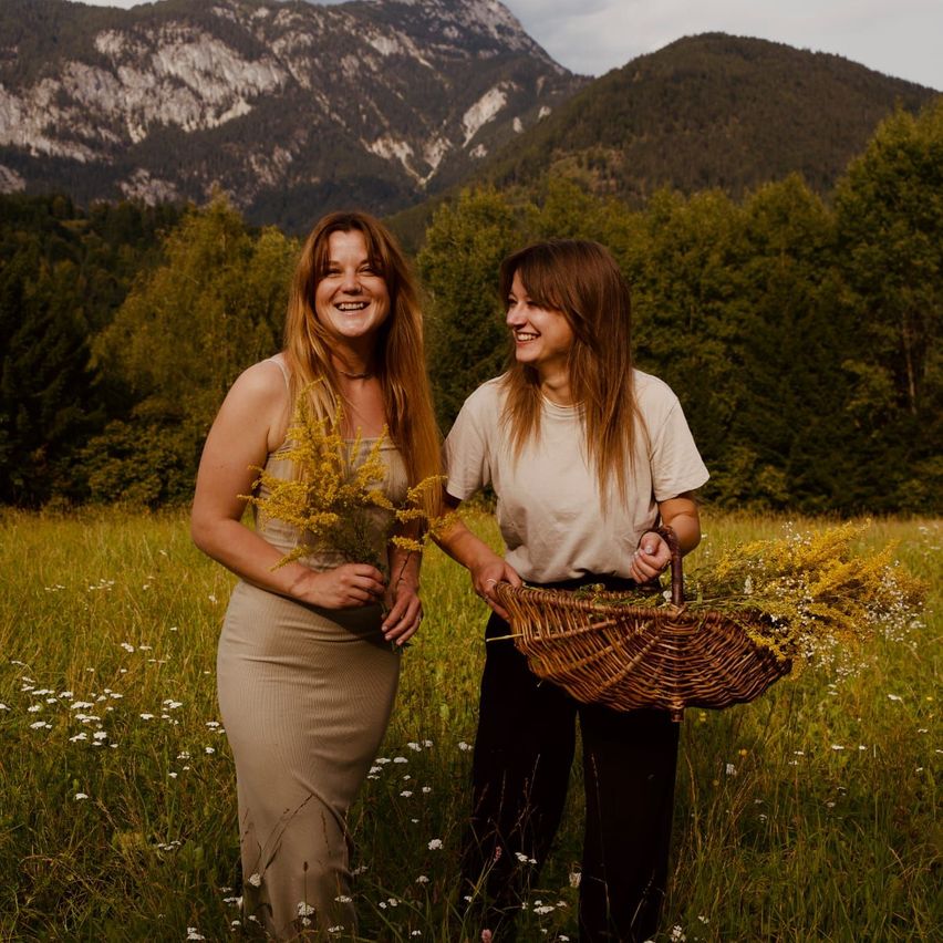 Zwei Frauen stehen in einem Feld, lächeln und halten einen Korb mit gelben Blumen. Sie sind von hohem Gras und Bäumen umgeben, mit Bergen im Hintergrund.